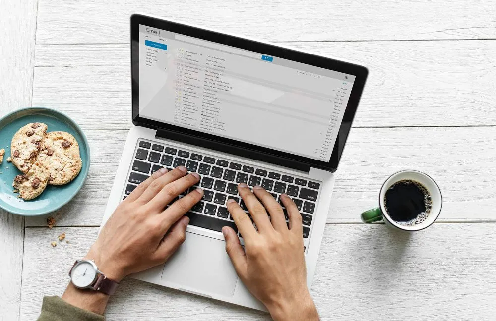 Overhead view of hands typing on a laptop showing an email inbox, beside a coffee cup and a plate of biscuits